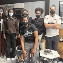 Group of six people wearing masks, standing indoors with tables and chairs nearby.