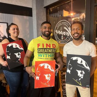 Three people holding portraits of a man with sunglasses and a hat, smiling inside a cafe or restaurant.