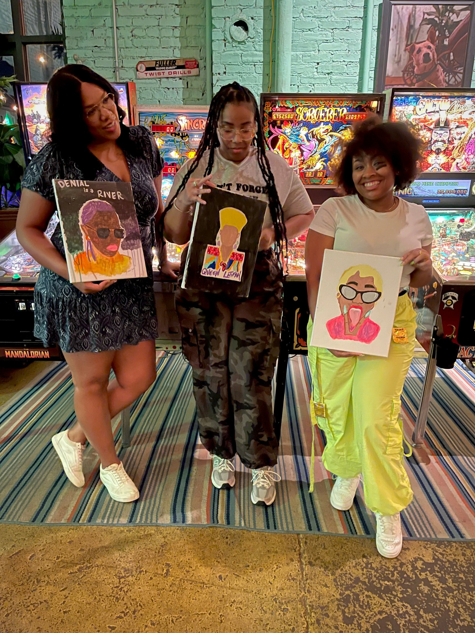 Three young women holding colorful portrait drawings in arcade with pinball machines in background.