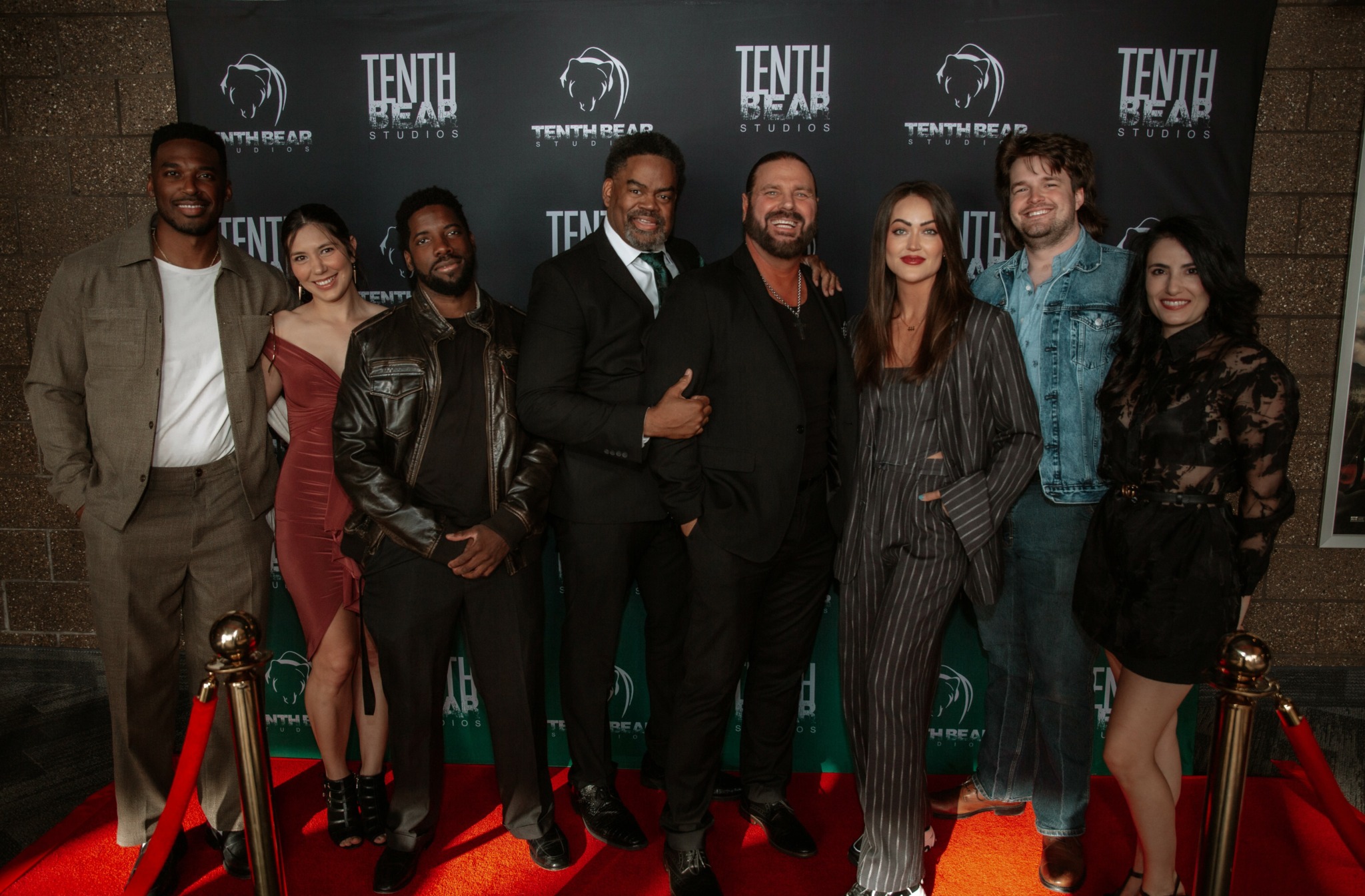 Group of eight people standing on red carpet in front of a black backdrop with event logos, smiling for photo.