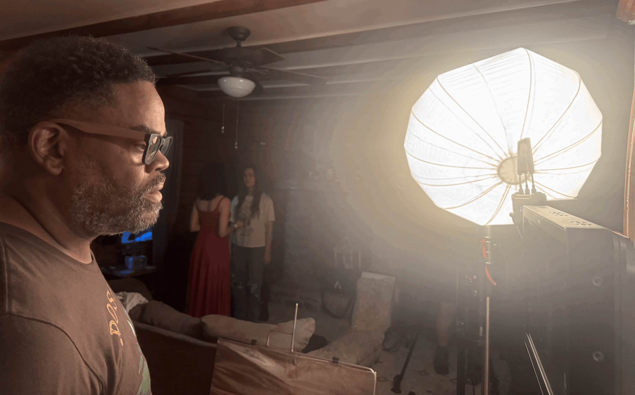 Man with glasses and beard in profile, indoors, near a bright studio light, with two women in background.