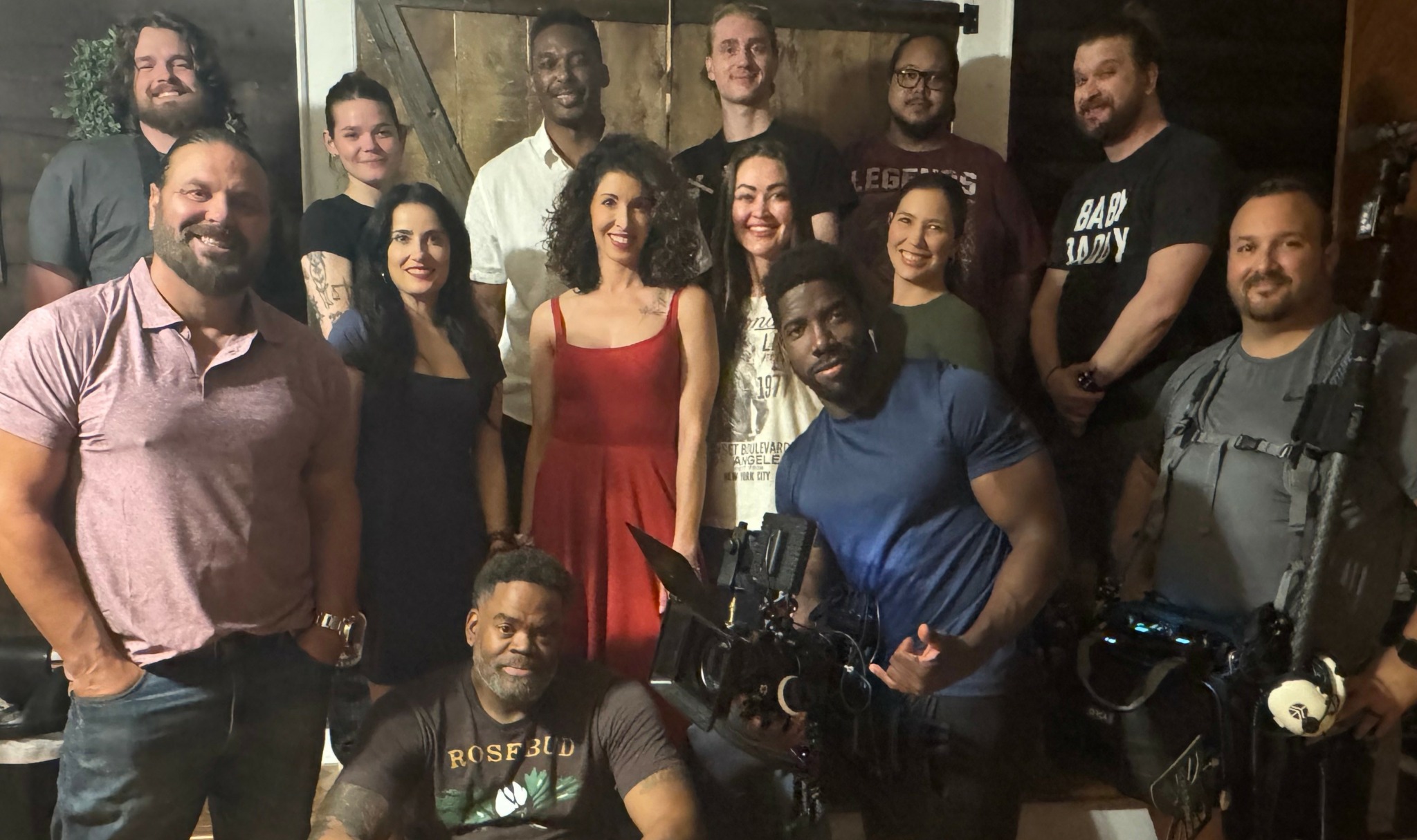 Group of 14 people indoors, some holding cameras, smiling, diverse in appearance, casual clothing, wooden background.