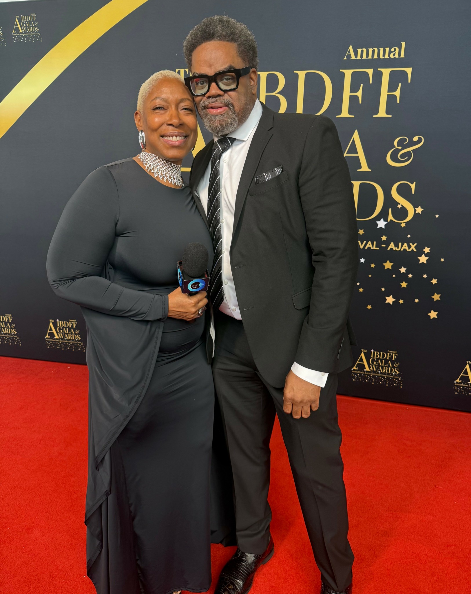 Two people in formal attire standing on red carpet at an event, smiling, with a backdrop displaying text and stars.