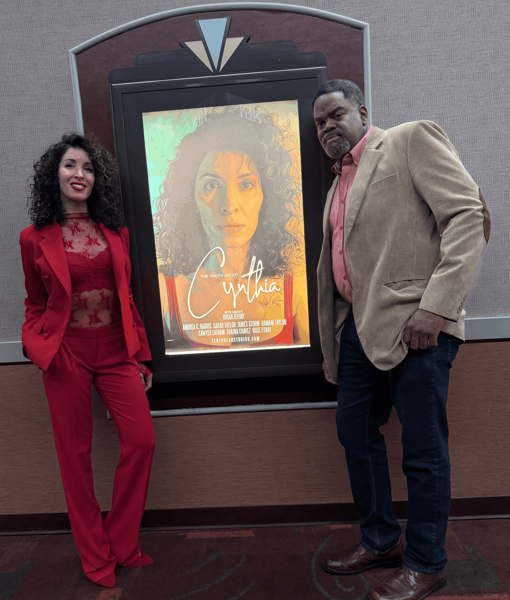 Two people standing next to a large movie poster display, smiling, in an indoor setting.