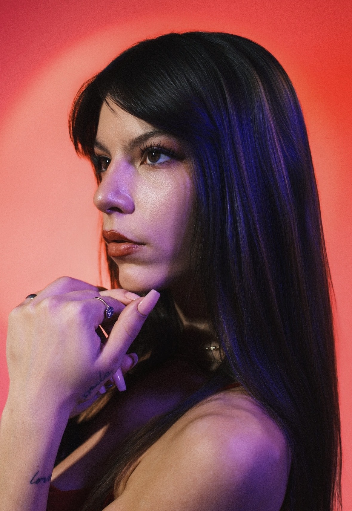 Woman with long dark hair and purple lighting, looking thoughtfully to the side, hand near chin.