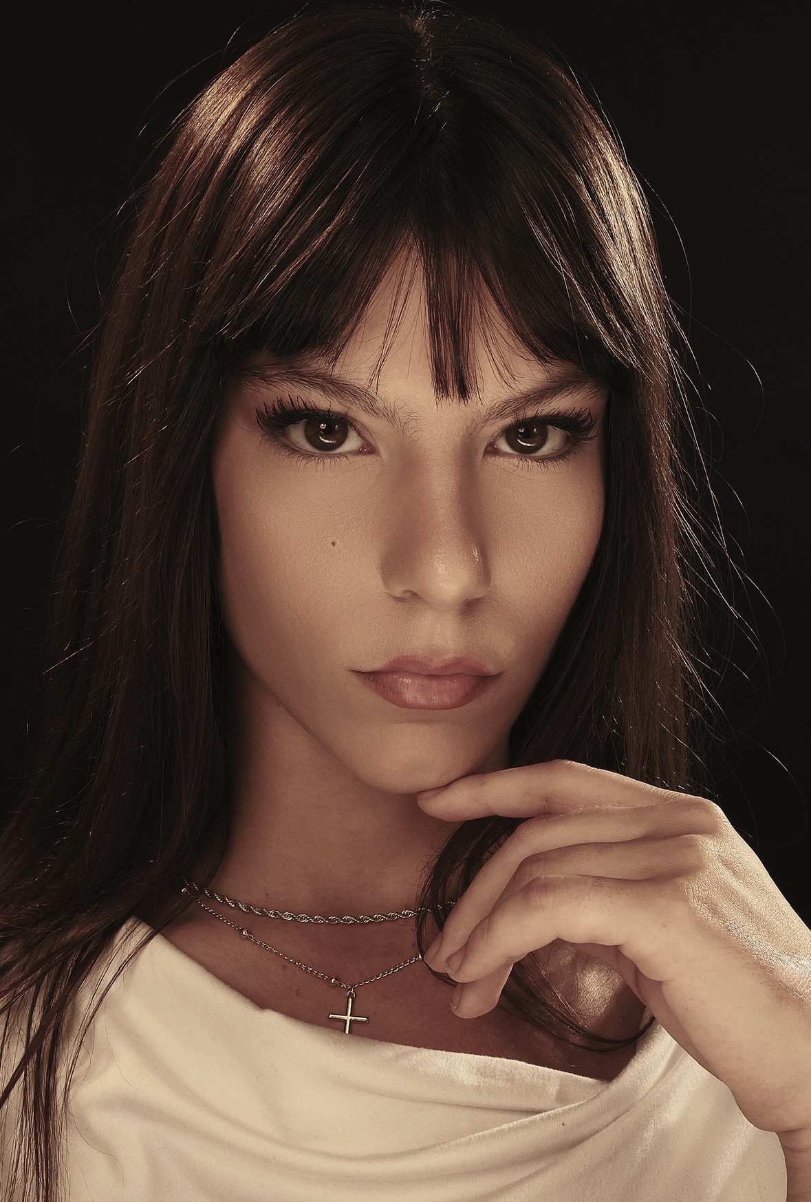 Close-up of a woman with long dark hair, wearing a white top and a cross necklace, looking at the camera.
