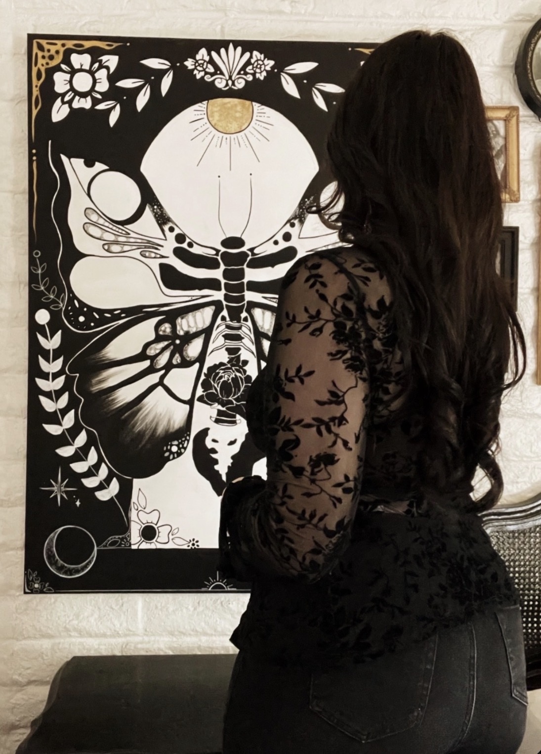 Woman with long dark hair wearing a black lace top, facing a butterfly and floral artwork on a wall.