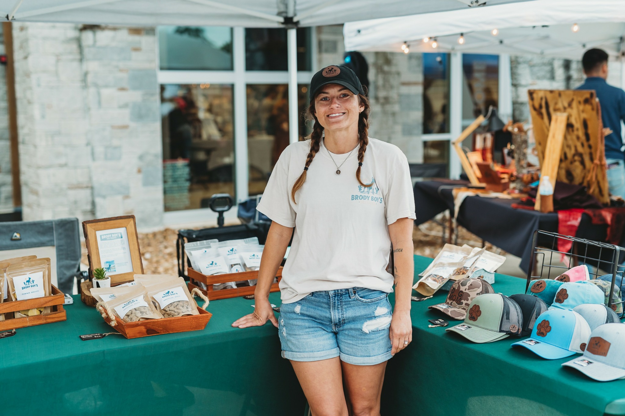 Smiling woman in white t-shirt, denim shorts, and cap standing at outdoor market stall with hats and framed pictures.