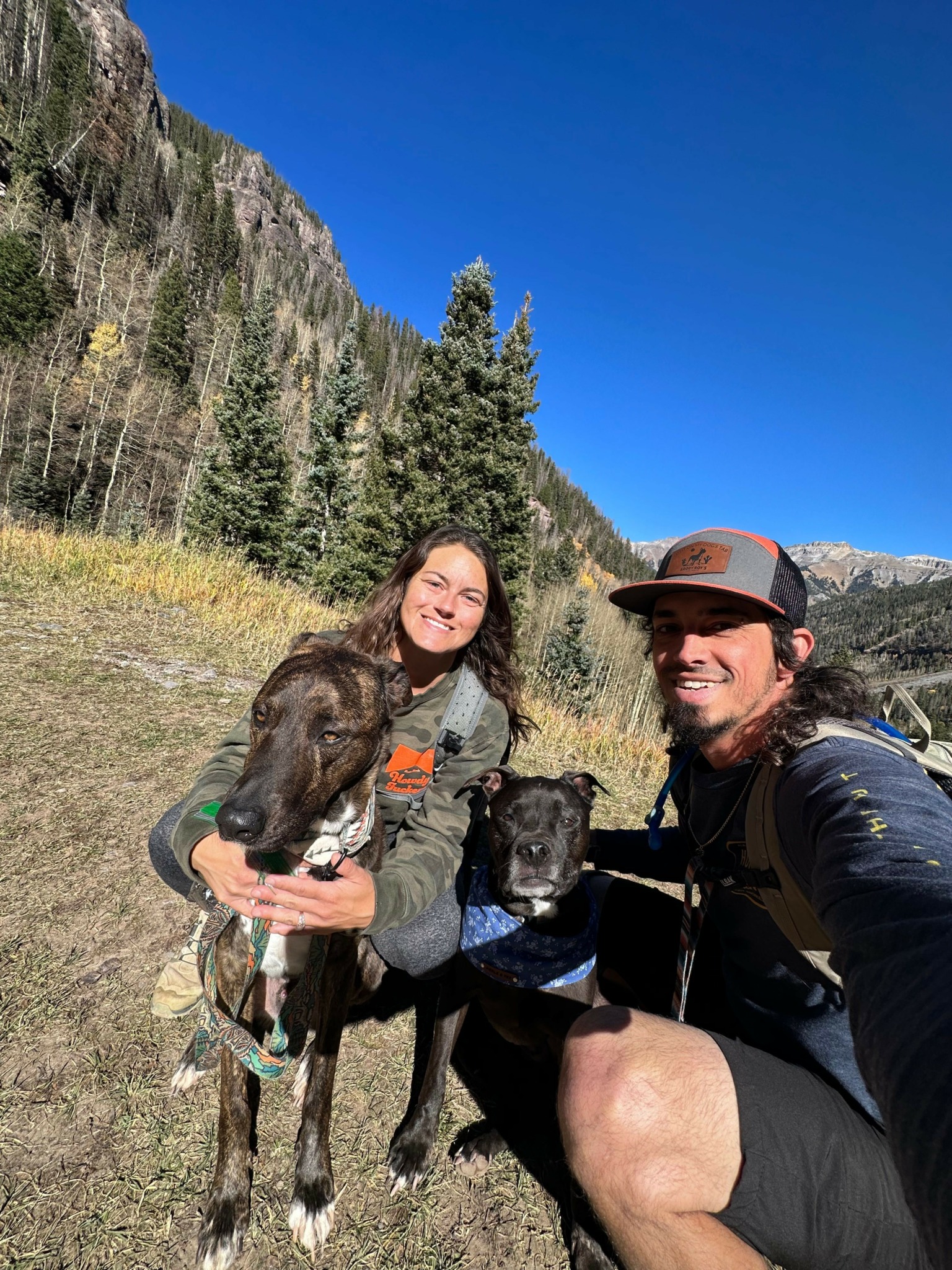 Two people outdoors with a dog, trees, and a clear blue sky in the background.