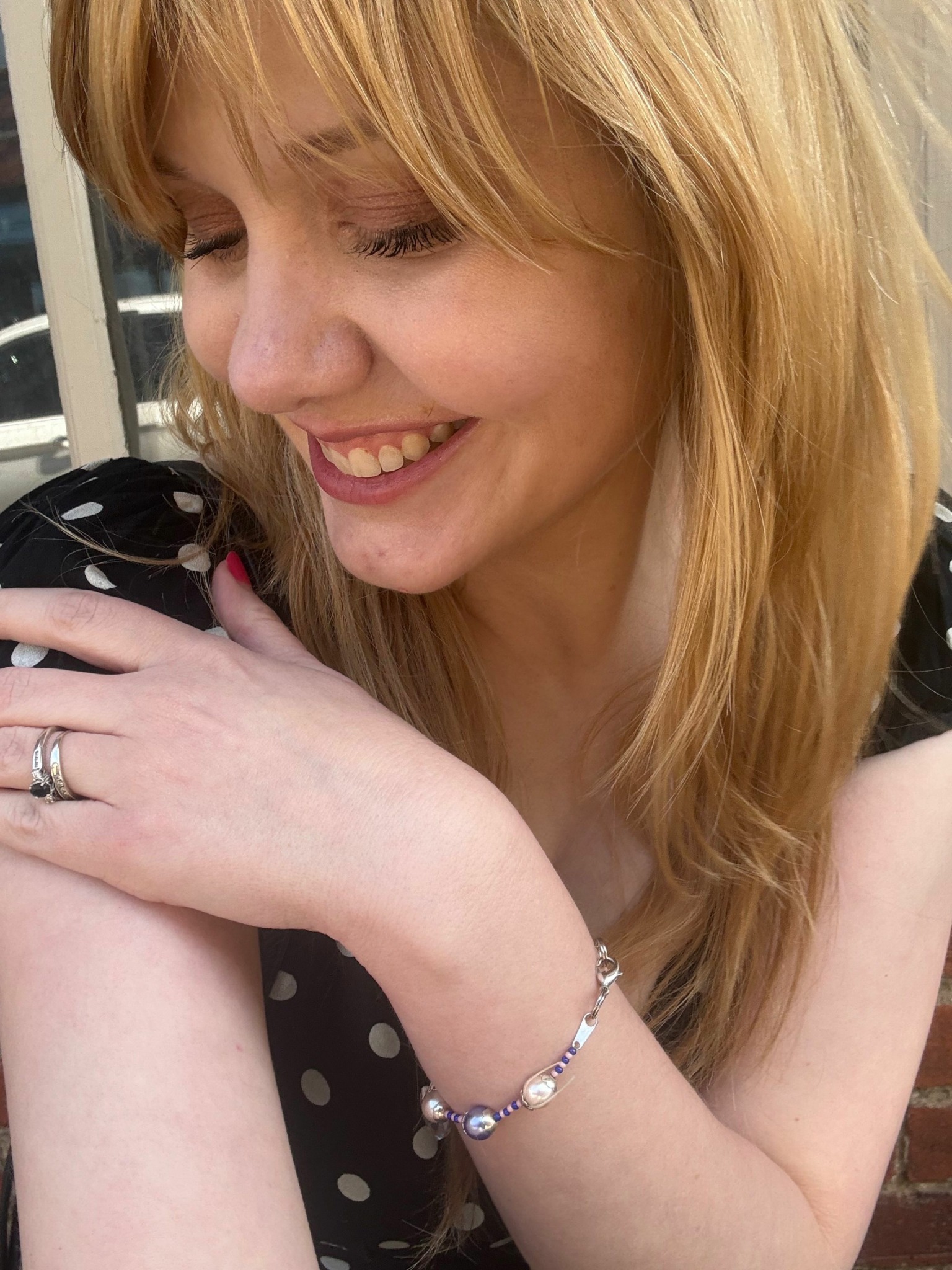 Woman with blonde hair smiling, resting her head on her arm, wearing a bracelet and ring, in a polka dot top.