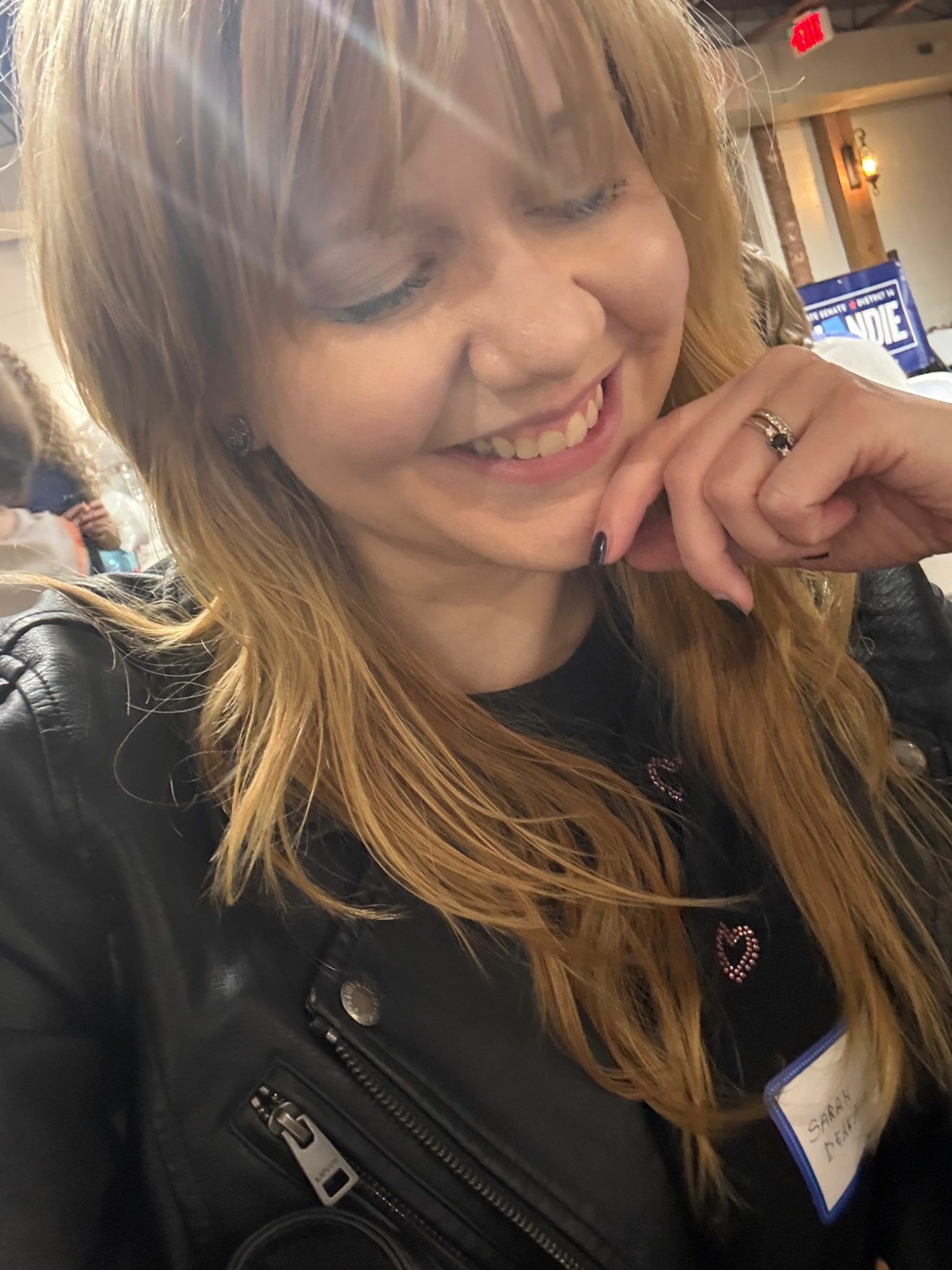 Young woman with long blonde hair smiling, holding her hand near her face, in an indoor setting.