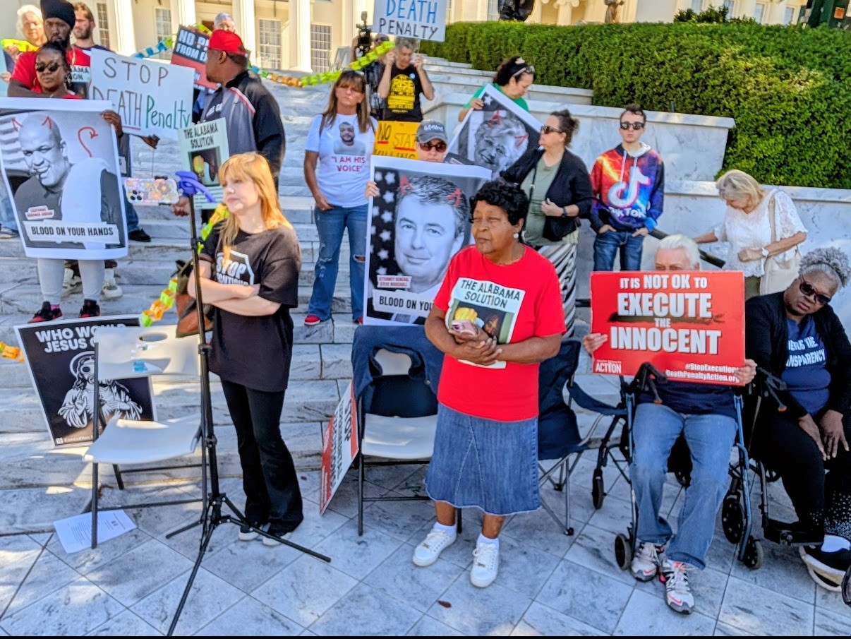 Group of people holding signs and banners in a public square, some seated, with buildings and greenery in background.