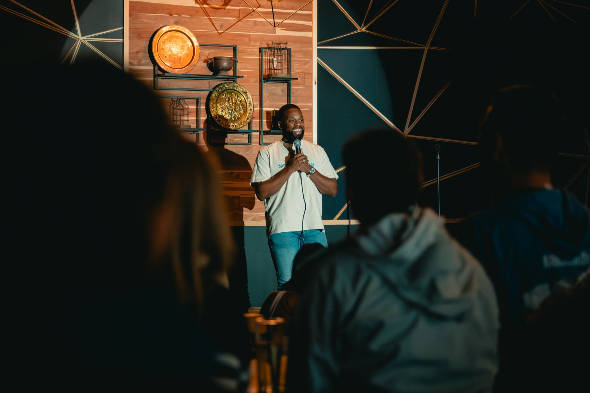 Person speaking on stage with audience in foreground, stage decorated with wooden panels and circular lights.