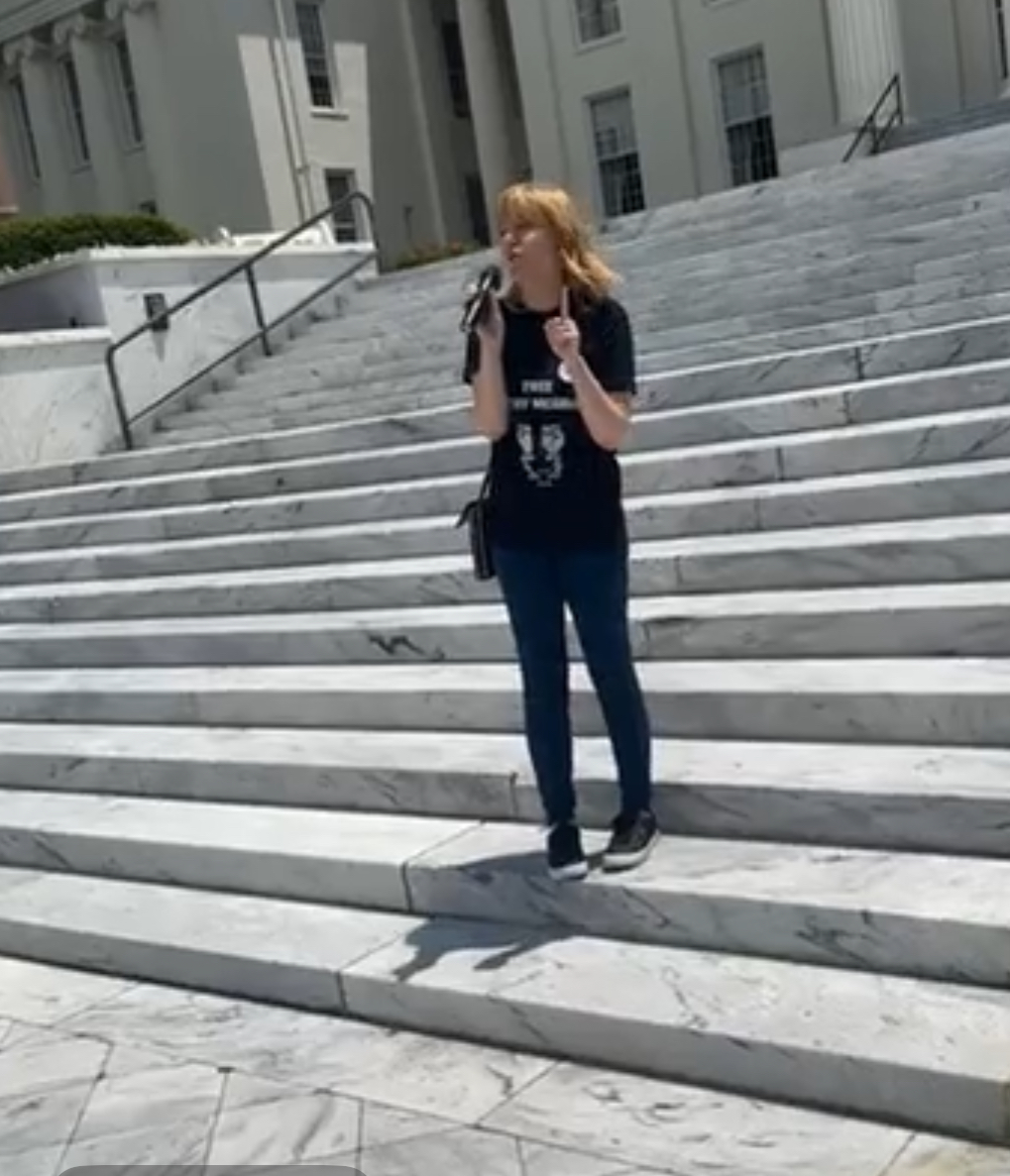 Person with long hair standing on marble stairs outside a building, holding a bag and wearing a dark shirt and jeans.