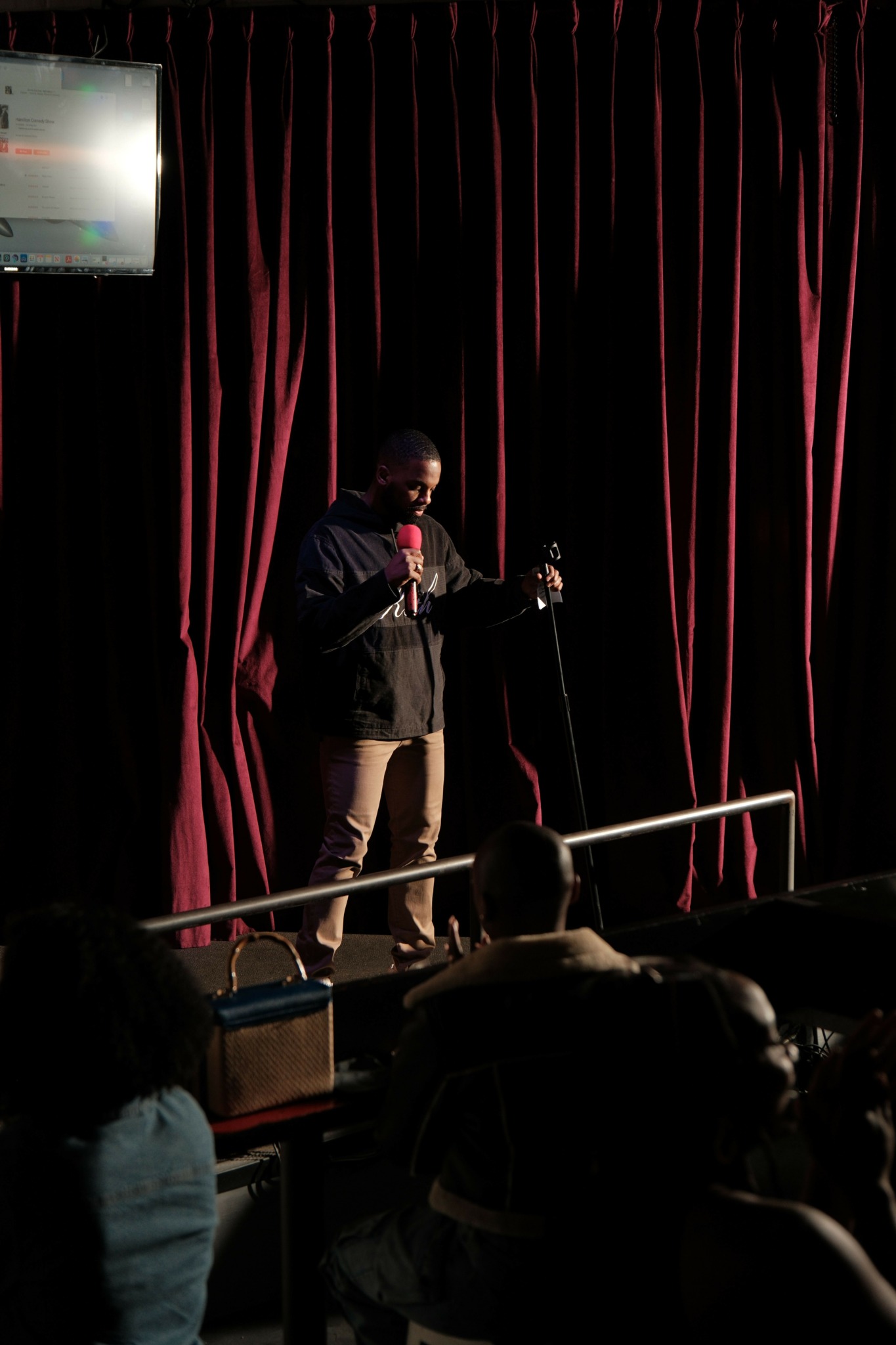 Person standing on stage with a microphone, facing an audience, in front of a red curtain.