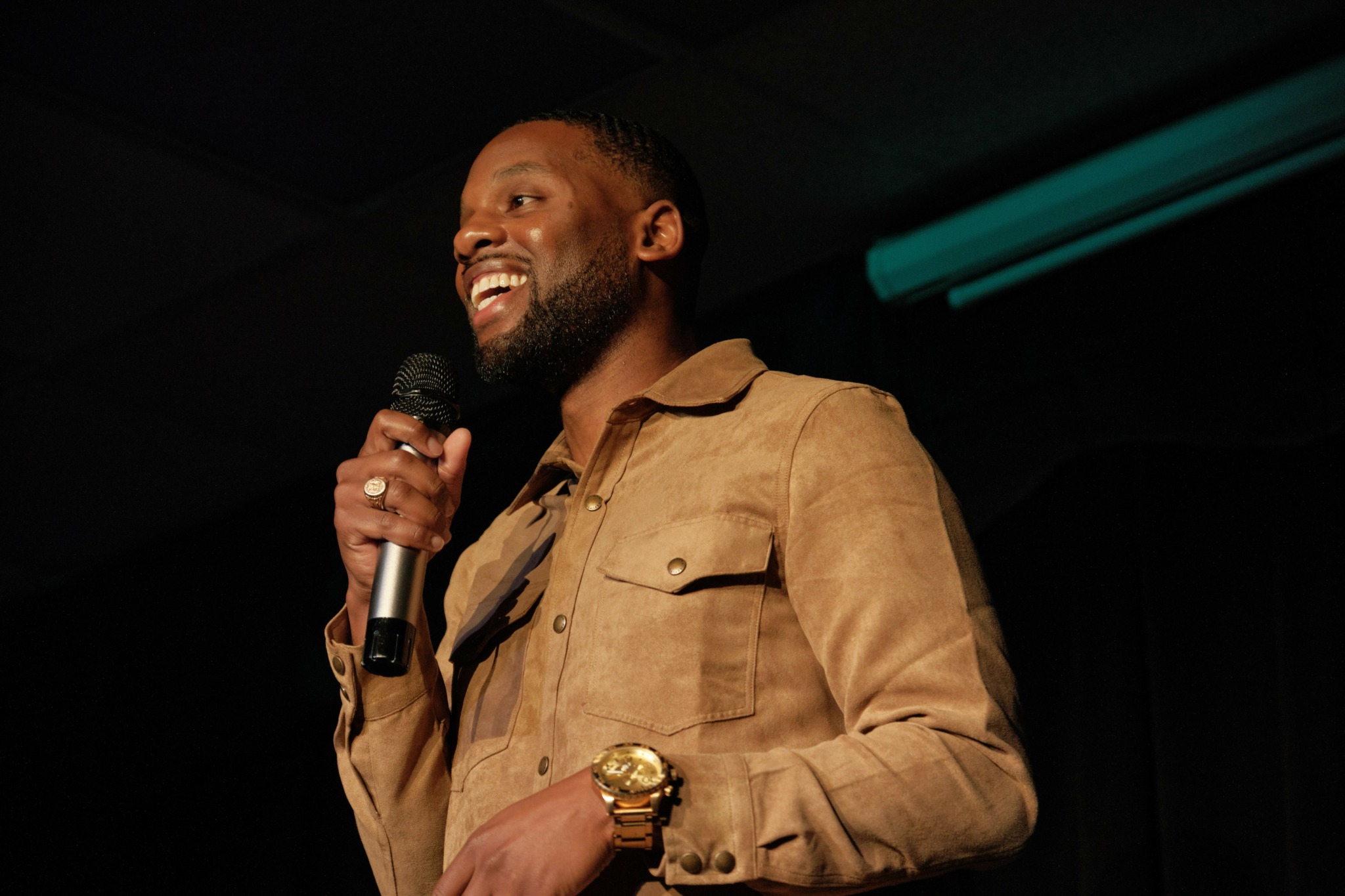 Man smiling and speaking into a microphone on stage, wearing a tan jacket and gold watch.