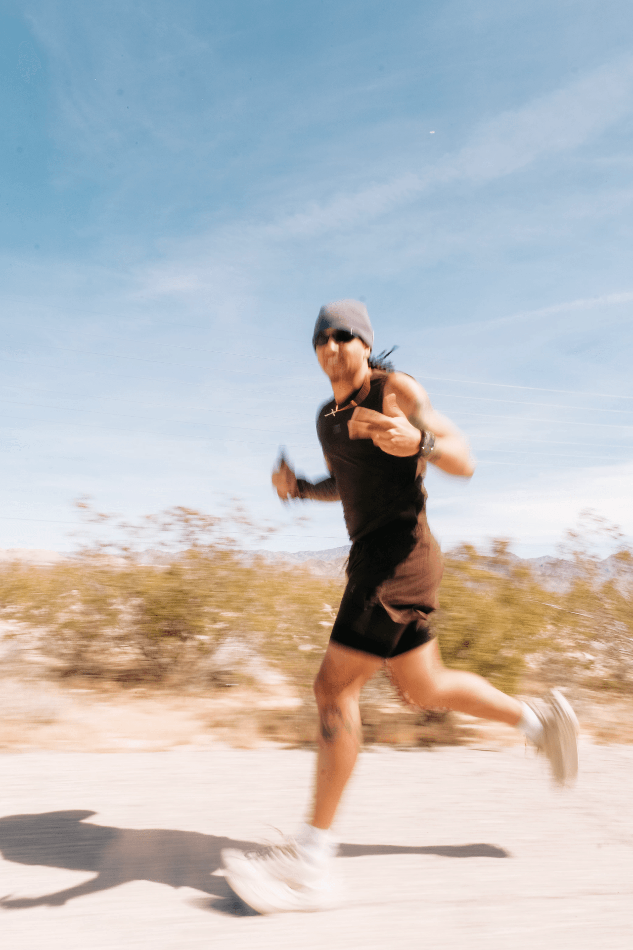 Person running outdoors wearing sunglasses, a hat, black tank top, and shorts, with a blue sky background.