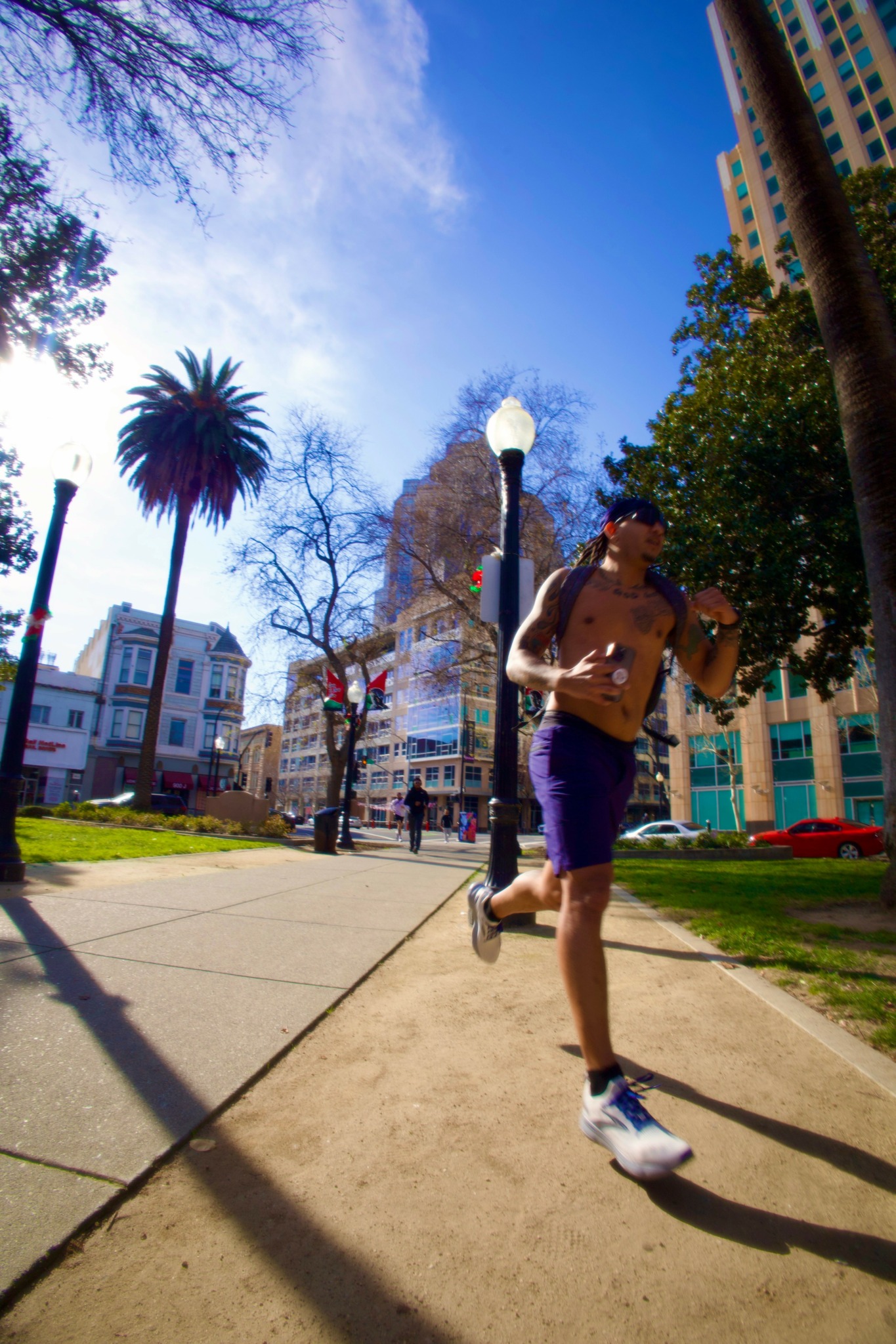 Person jogging on a sidewalk in a city park with trees, buildings, and a blue sky in the background.