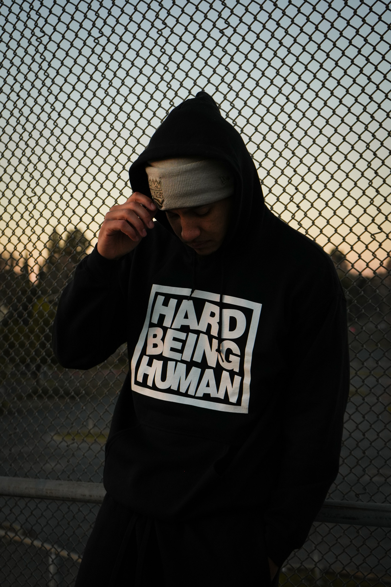 Person wearing a hoodie and cap, standing in front of a chain-link fence during sunset.