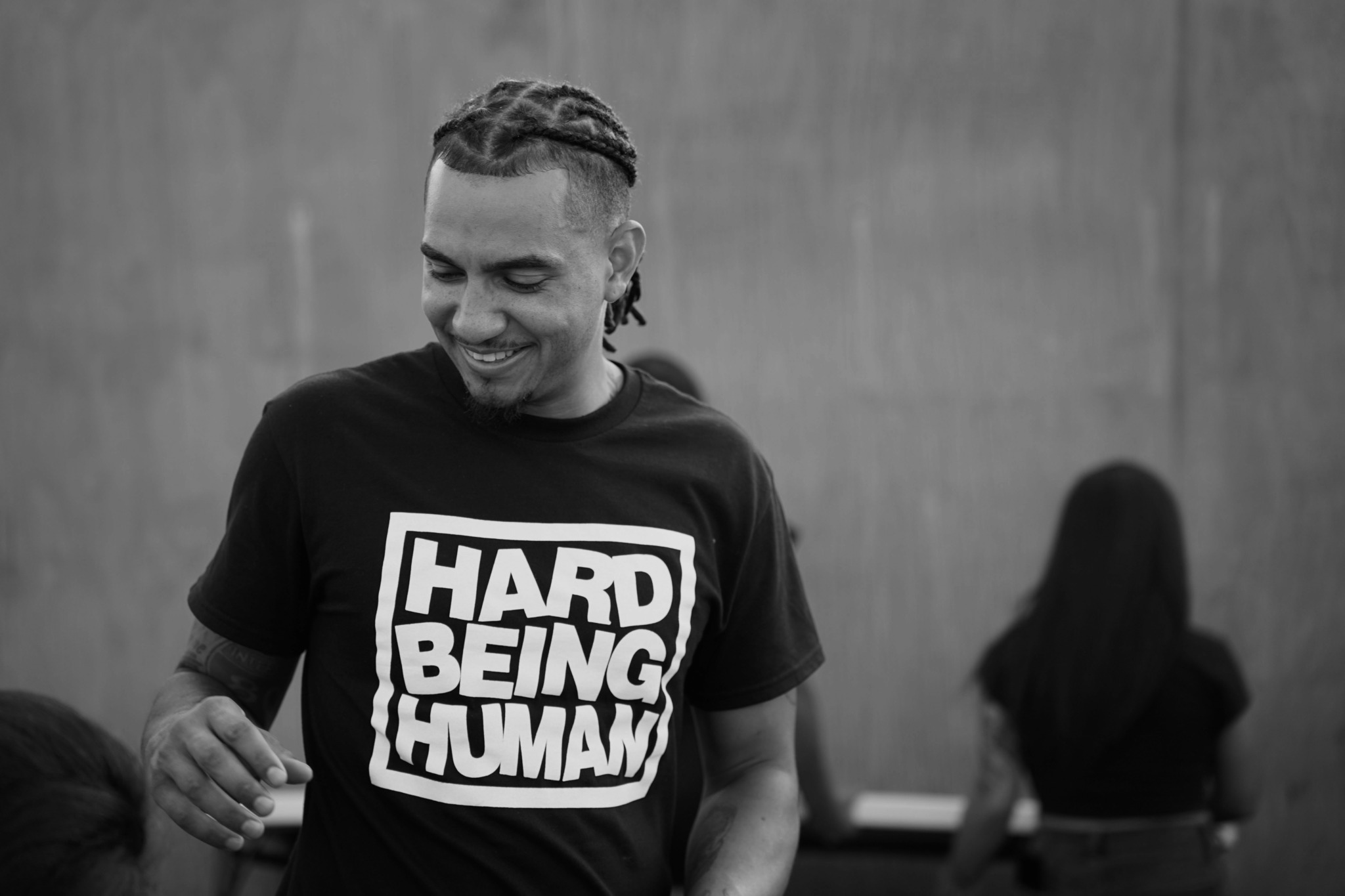 Smiling person with braided hair wearing a T-shirt that says 'HARD BEING HUMAN' in front of a wall.