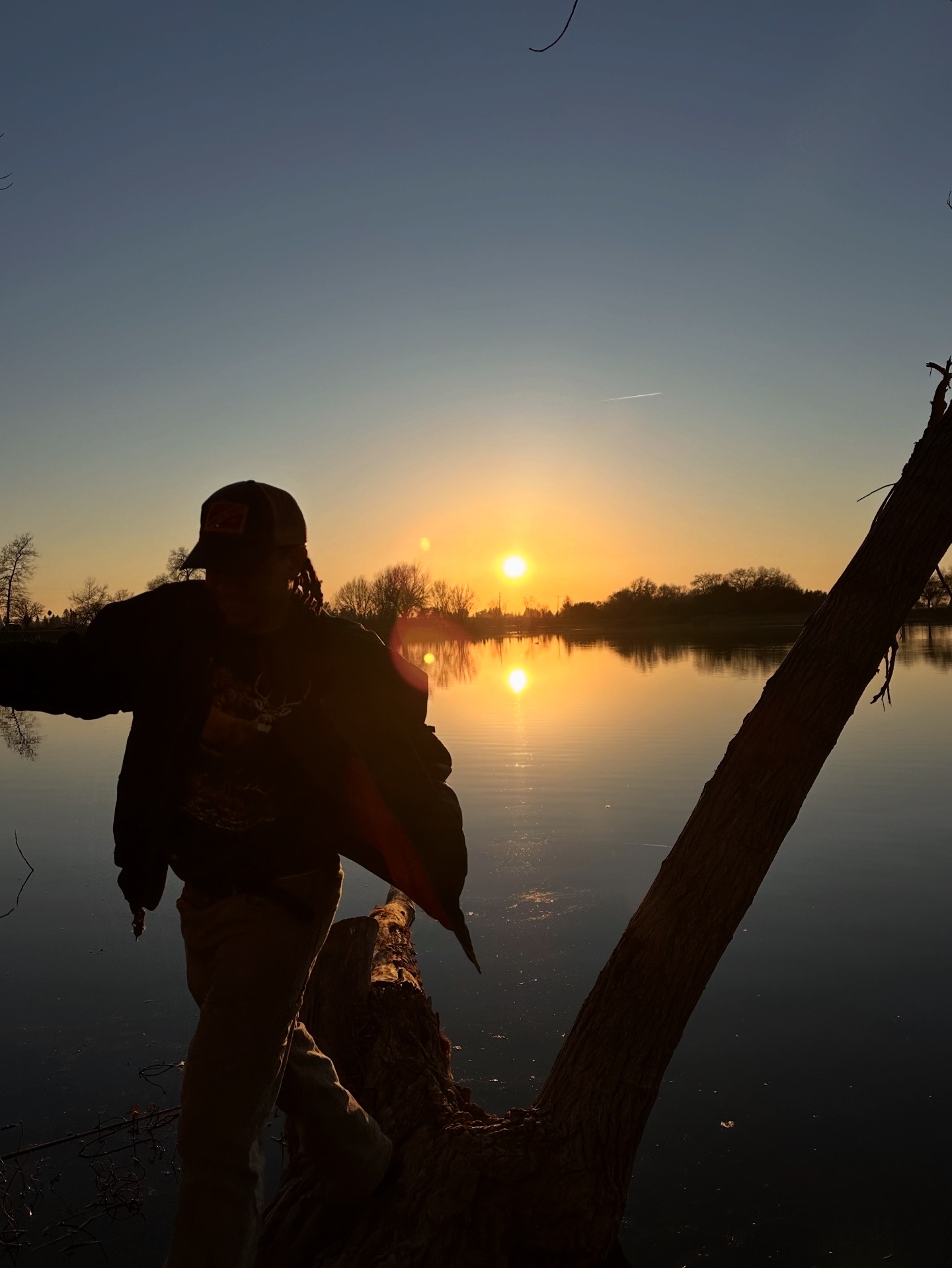 Person standing by a river at sunset with trees and water reflections.