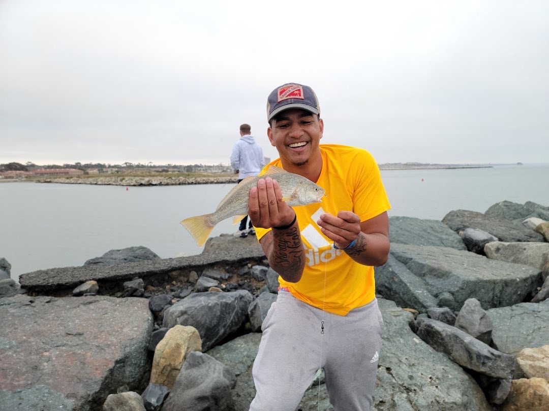 Person smiling, holding a fish near a rocky shoreline with water and cloudy sky in background.