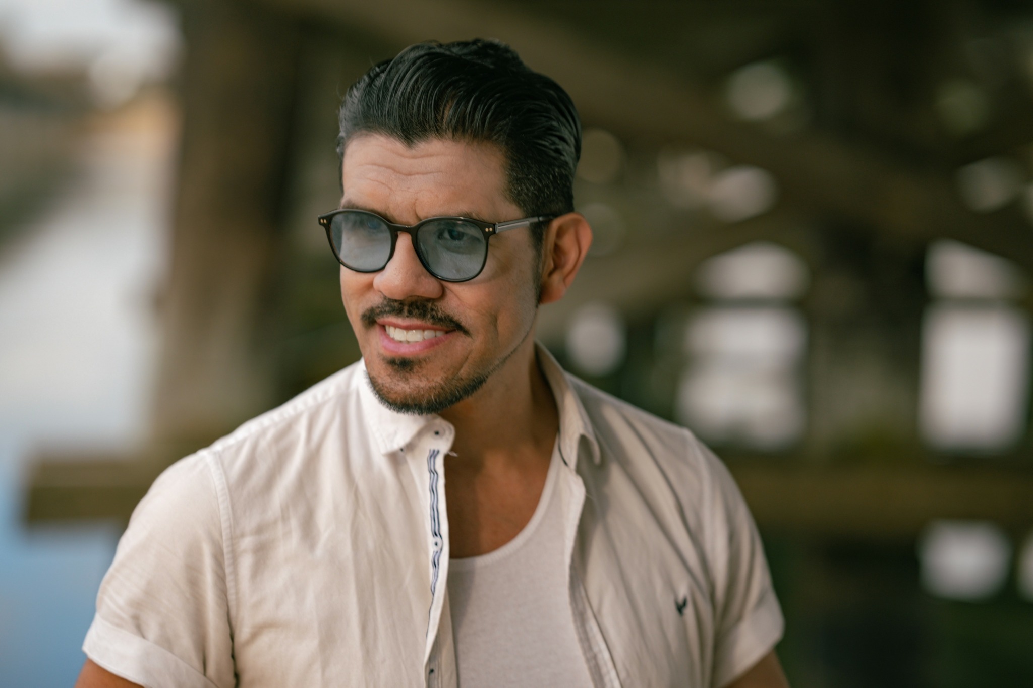 Man with dark hair, glasses, and a beard, smiling outdoors in sunlight, wearing a light-colored shirt.