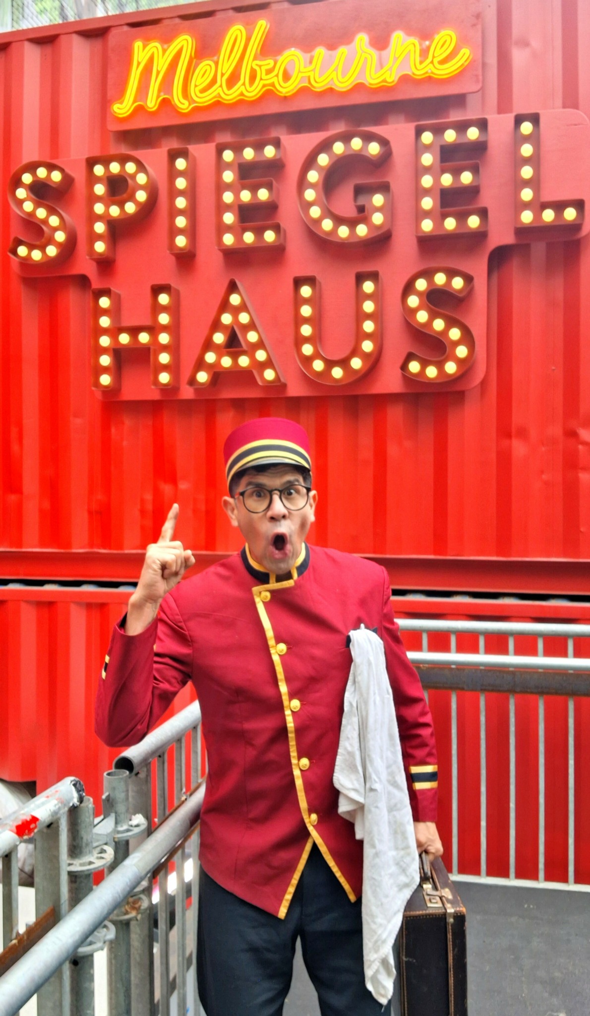 Person in red uniform with glasses and a hat, holding a white cloth, standing in front of a red wall with signs.