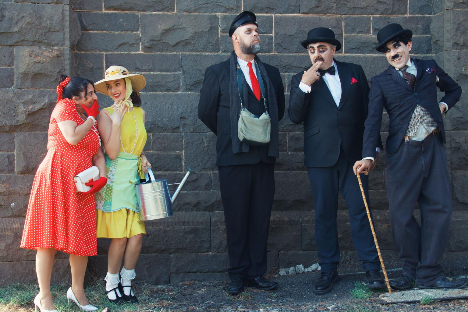 Four people dressed in vintage clothing standing against a brick wall, two women on the left and two men on the right.
