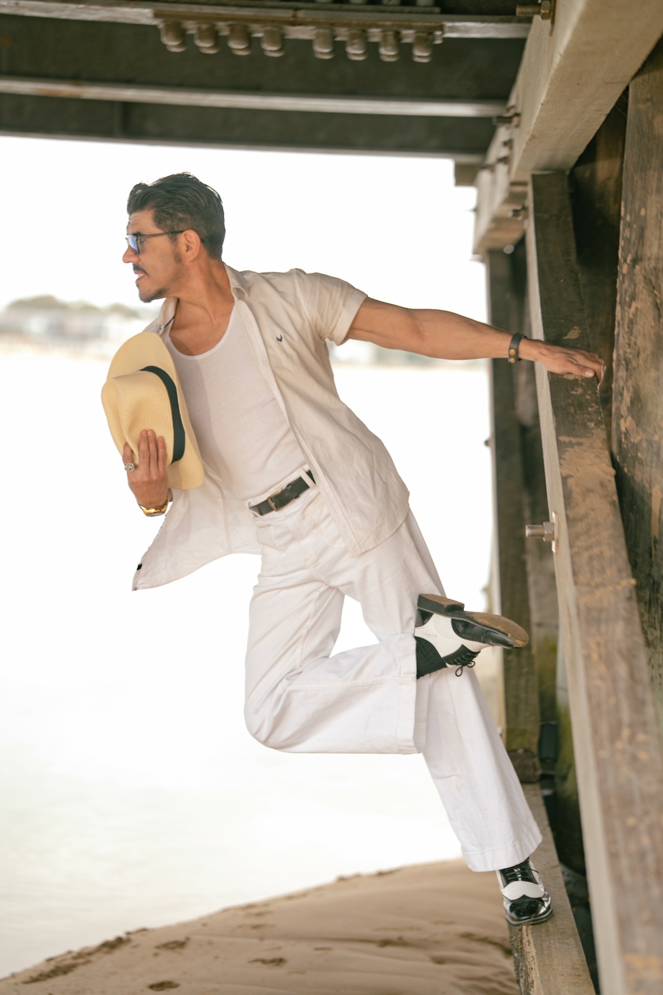 Man in white clothes jumping under a bridge holding a hat, with a sandy ground and bright background.