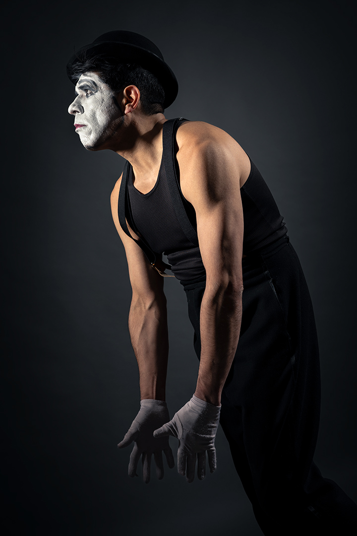 Performer with white face paint, black hat, black sleeveless top, and gloves, leaning forward against a dark background.