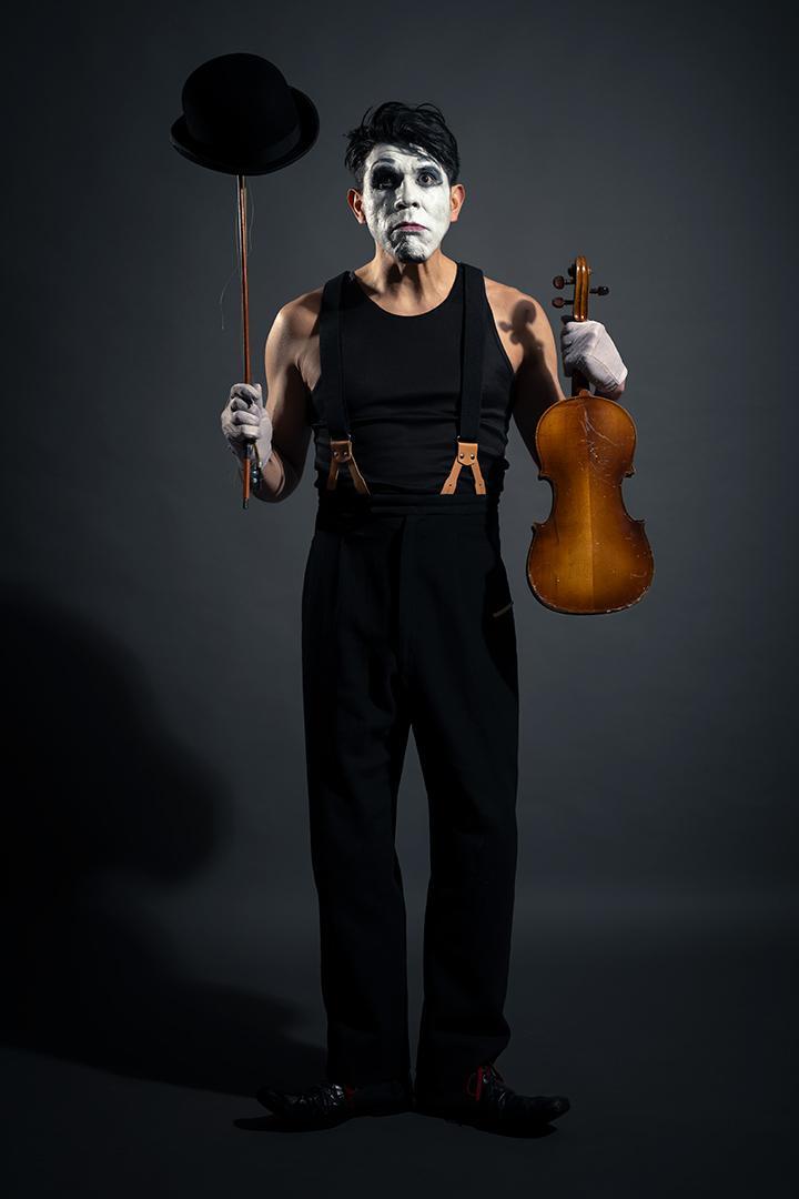 Person with white face paint, black hat, holding a violin and a hat on a stick, standing against a dark background.