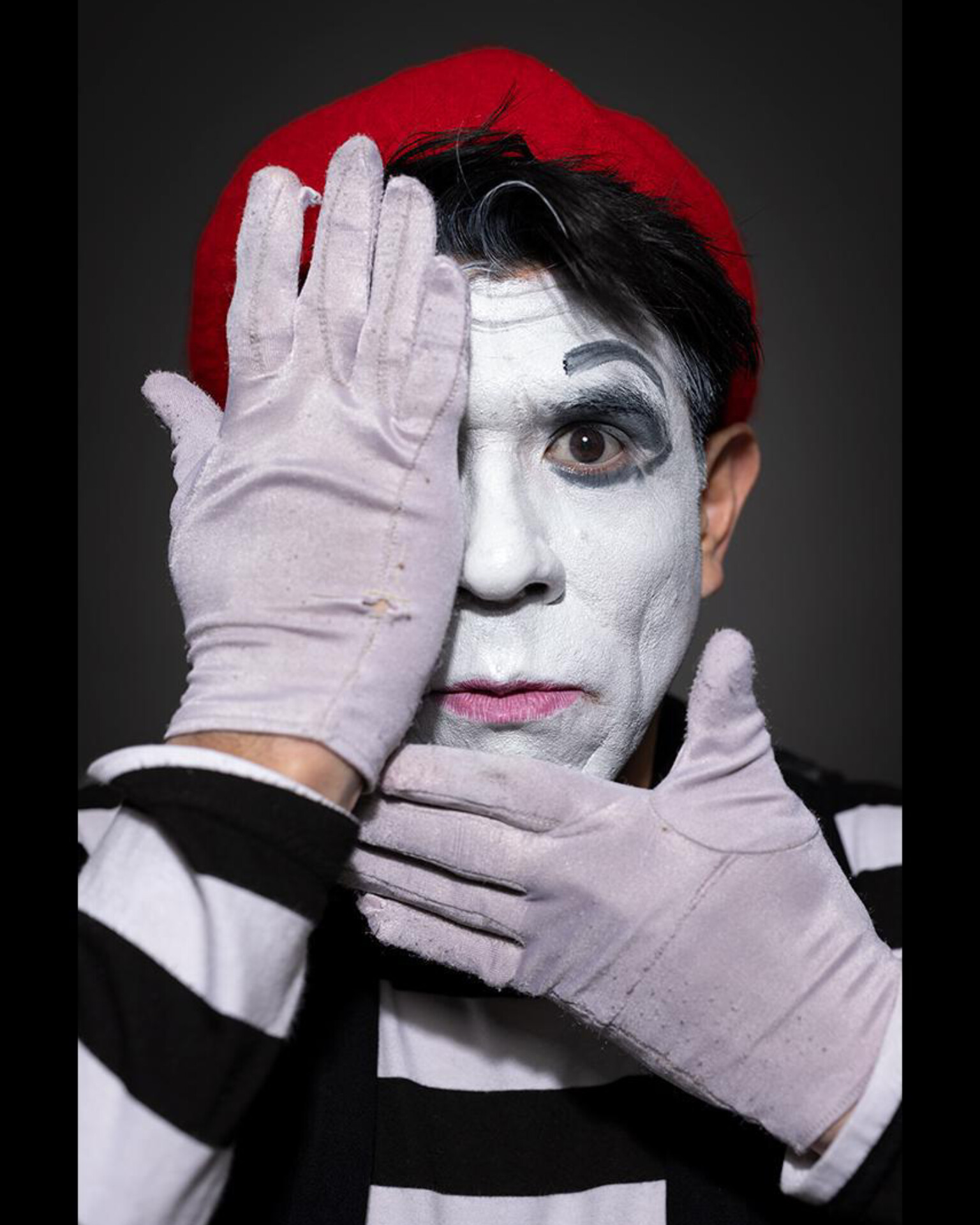 Person with face painted white, wearing a red hat and striped shirt, covering one eye with gloved hand.