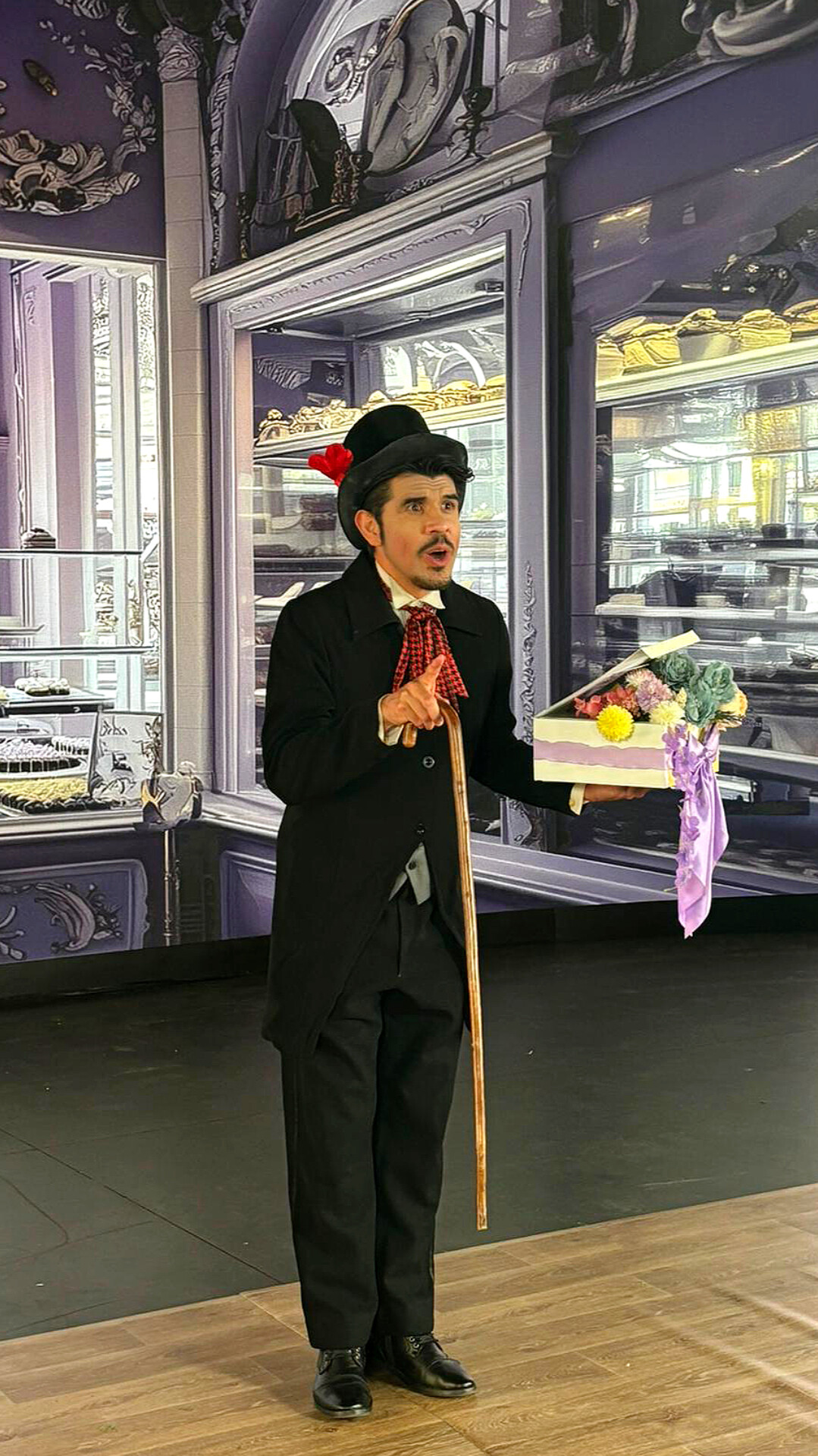 Man dressed in vintage costume with a cane, standing indoors near display cases with desserts and pastries.