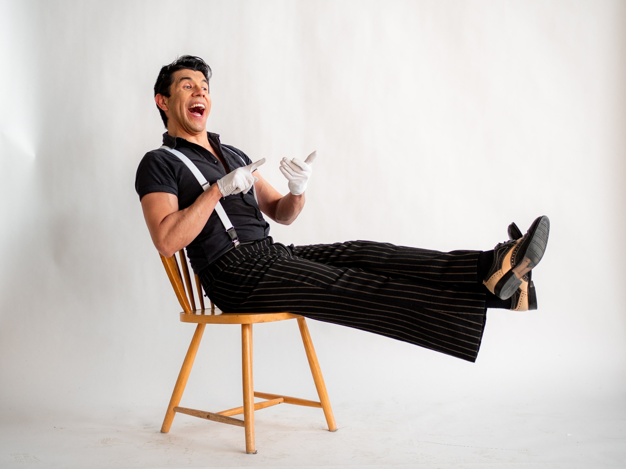 Person sitting on a chair with legs extended, laughing, wearing black striped pants and gloves, against a plain white background.