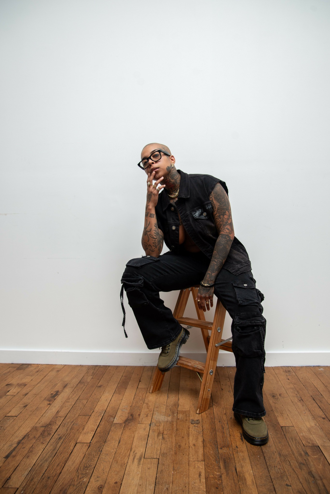 Person with tattoos and glasses sitting on a wooden stool against a plain white wall, with one hand on chin.