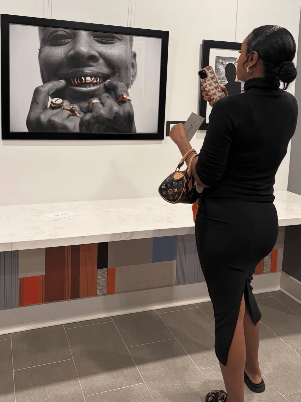 Woman in black dress with a handbag looking at a black-and-white photograph of a smiling person with gold teeth.