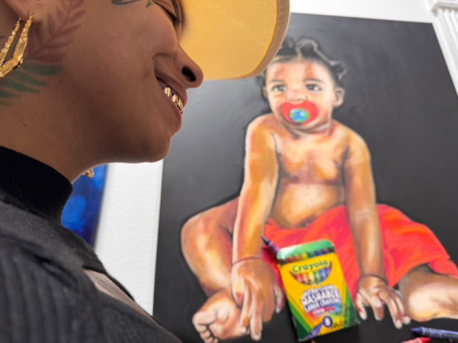Person smiling and looking at a colorful painting of a child with a pacifier, sitting on a red cloth.