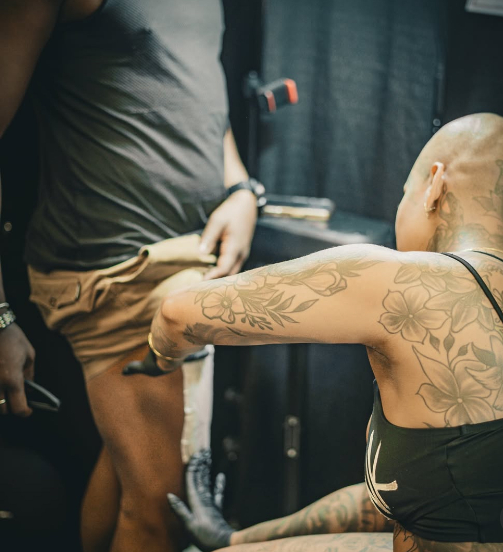 Tattooed person with a shaved head and black top reaching out, with another person in shorts nearby.