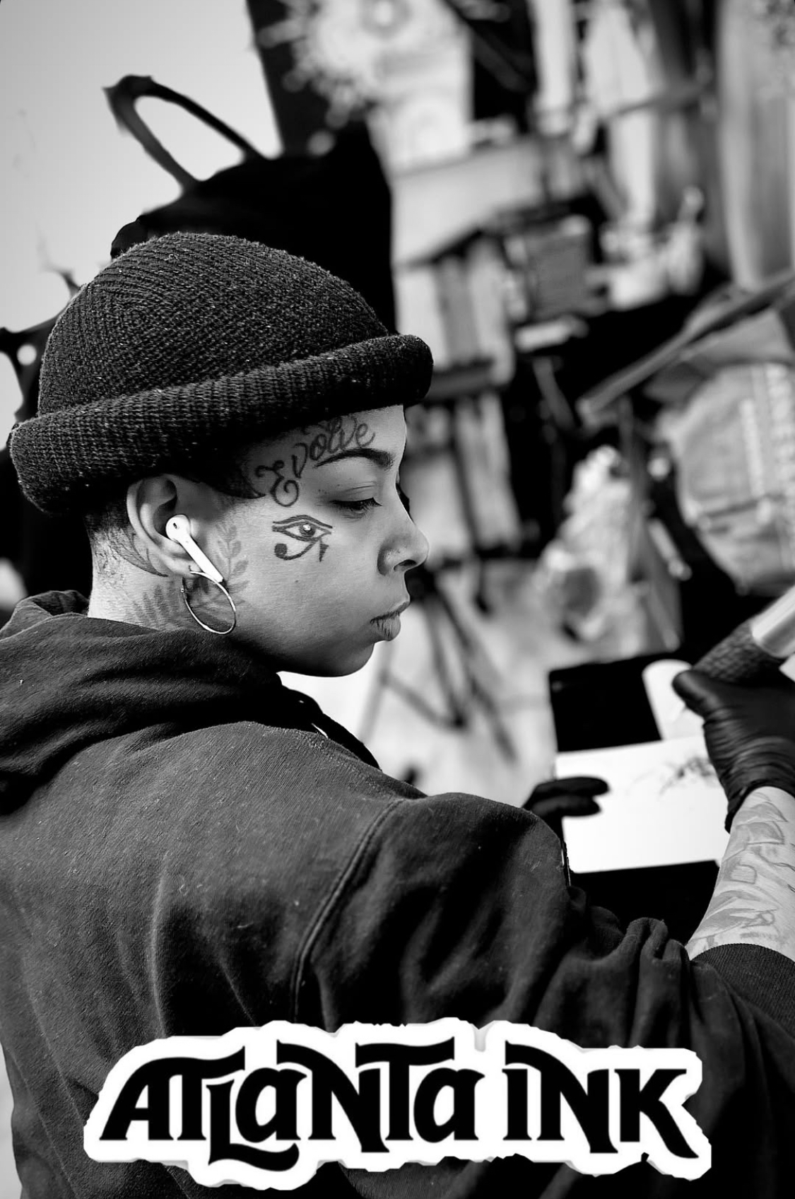 Person with tattoos, wearing a beanie, sitting at a table in a cluttered room.