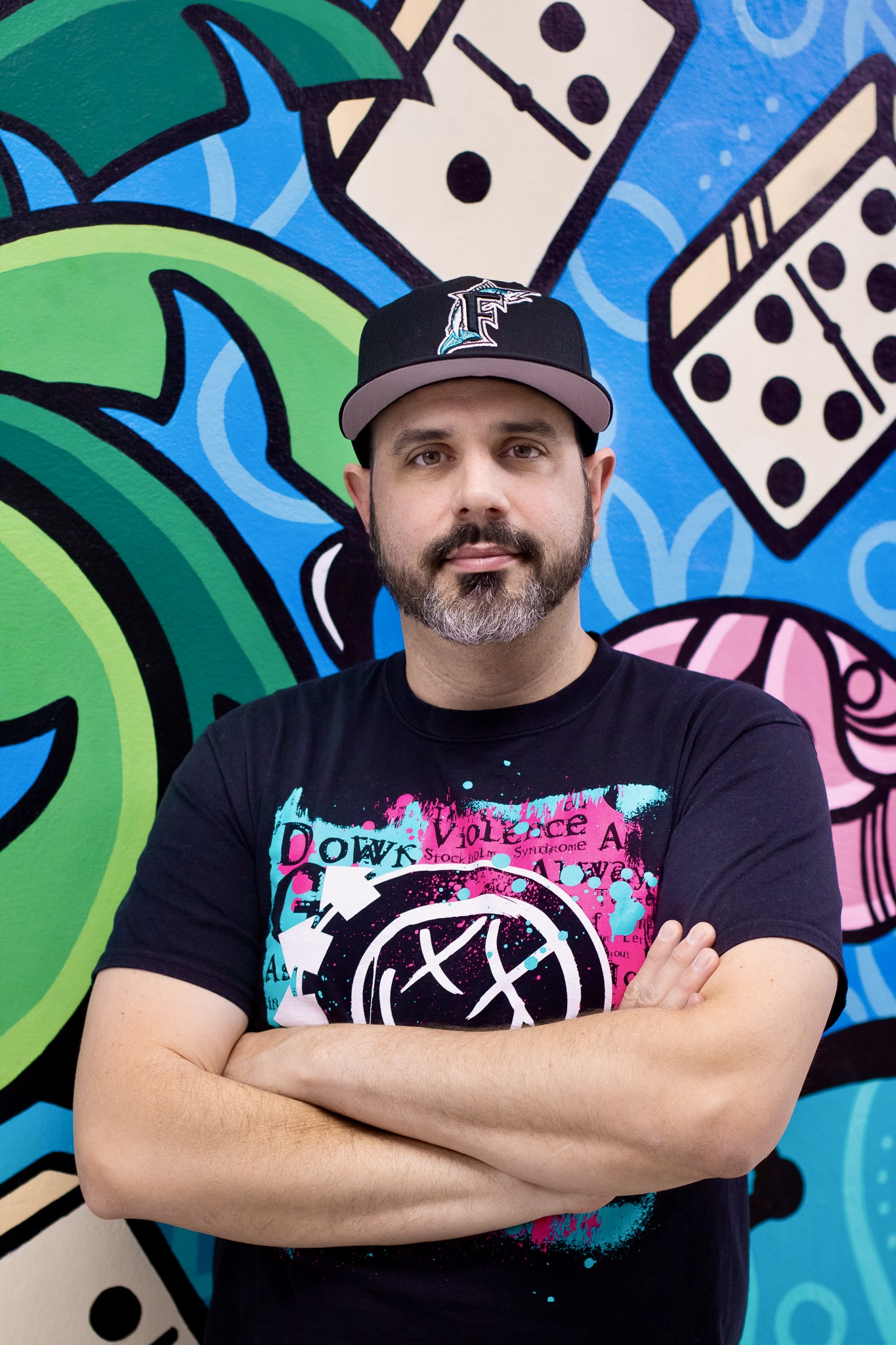 Man with beard wearing a black cap and T-shirt, standing with arms crossed in front of colorful graffiti background.