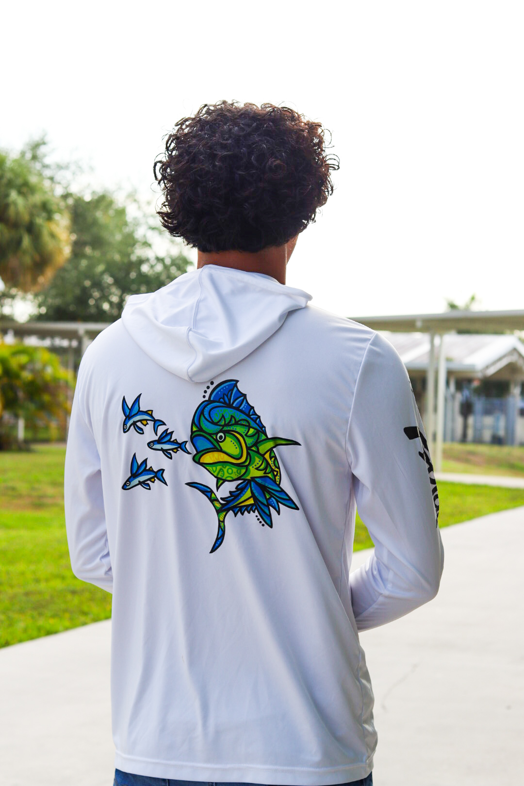 Person with curly hair wearing a white hoodie with colorful fish design on back, outdoors in a park-like setting.