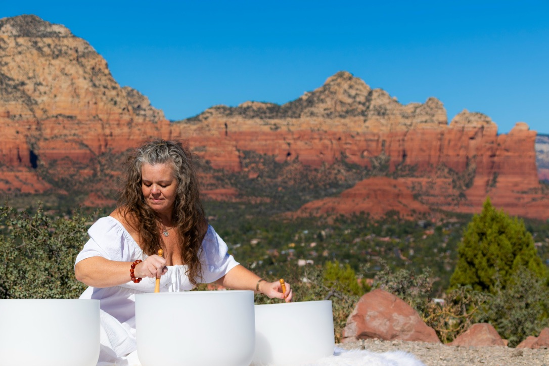 Woman with long hair in white shirt playing a large white drum outdoors with red rock formations in background.