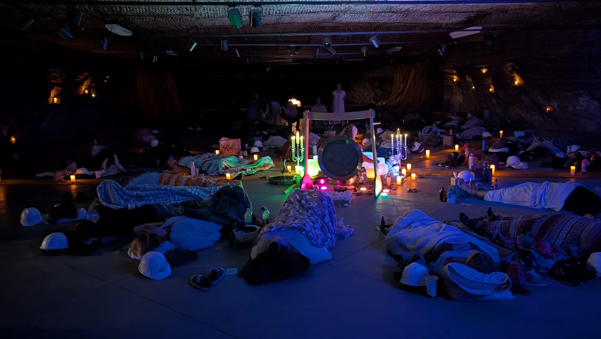 People lying on the floor in a dark room with candles and a central illuminated object.