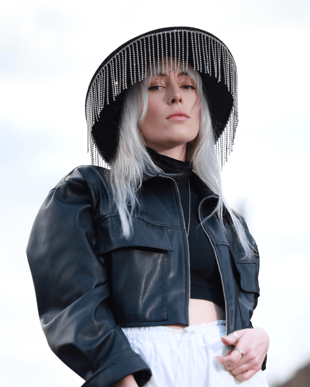 Woman wearing a large black hat with hanging beads, black leather jacket, and white pants, standing outdoors.