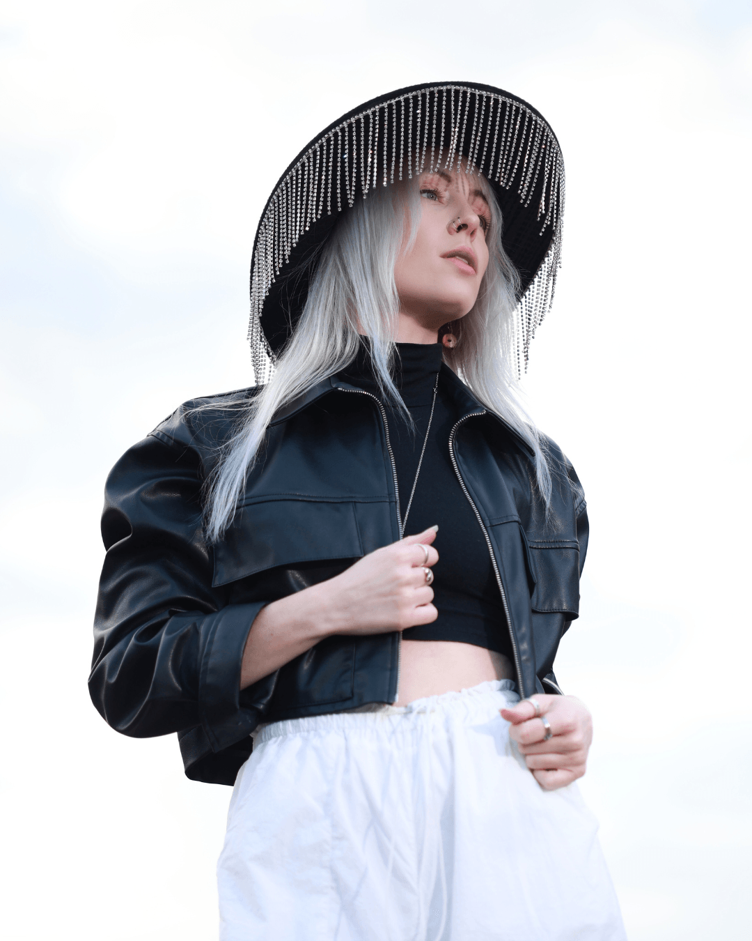Woman with long gray hair wearing a large black hat, black cropped jacket, and white pants outdoors.