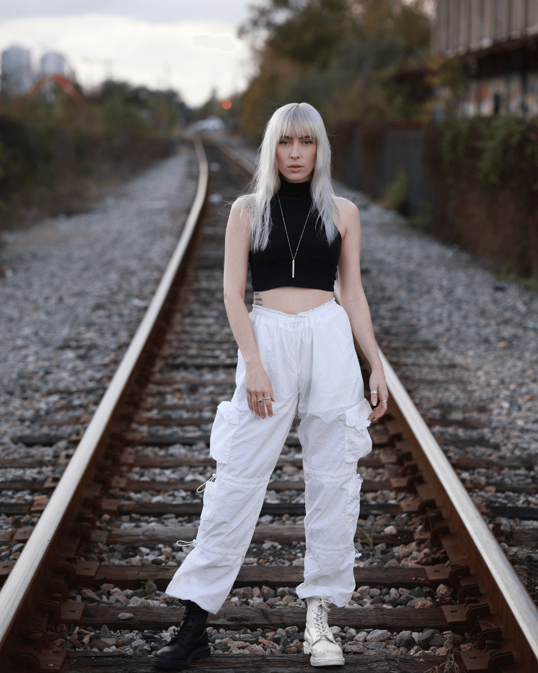 Young woman with long, straight, platinum blonde hair stands on railway tracks wearing a black sleeveless crop top and white baggy pants.