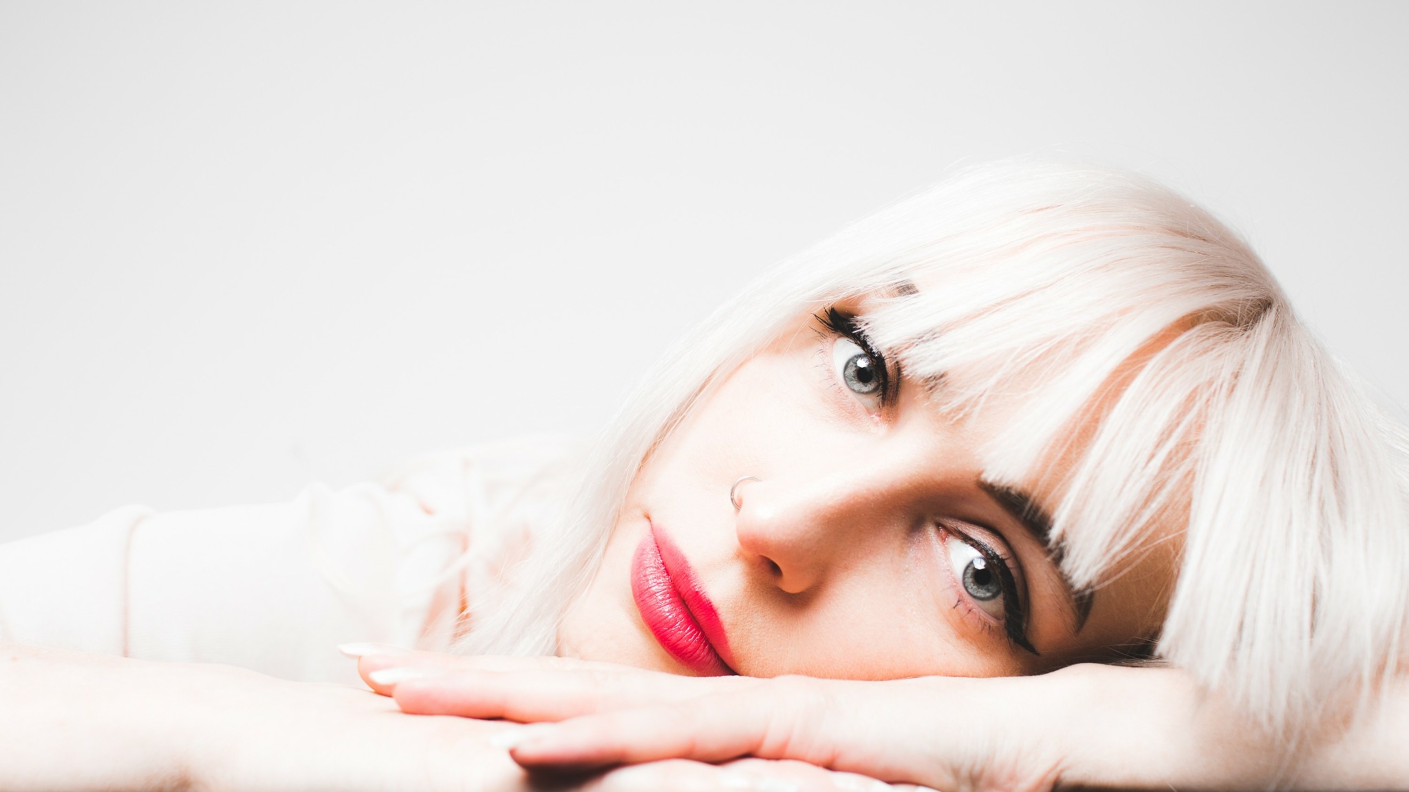Close-up of a woman with platinum blonde hair lying down, resting her head on her hands, with a neutral background.