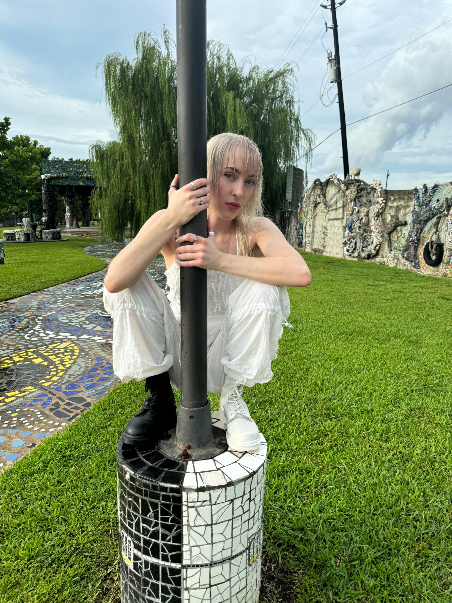 Person with light hair and white clothing crouching behind a pole outdoors, grassy area and trees in background.
