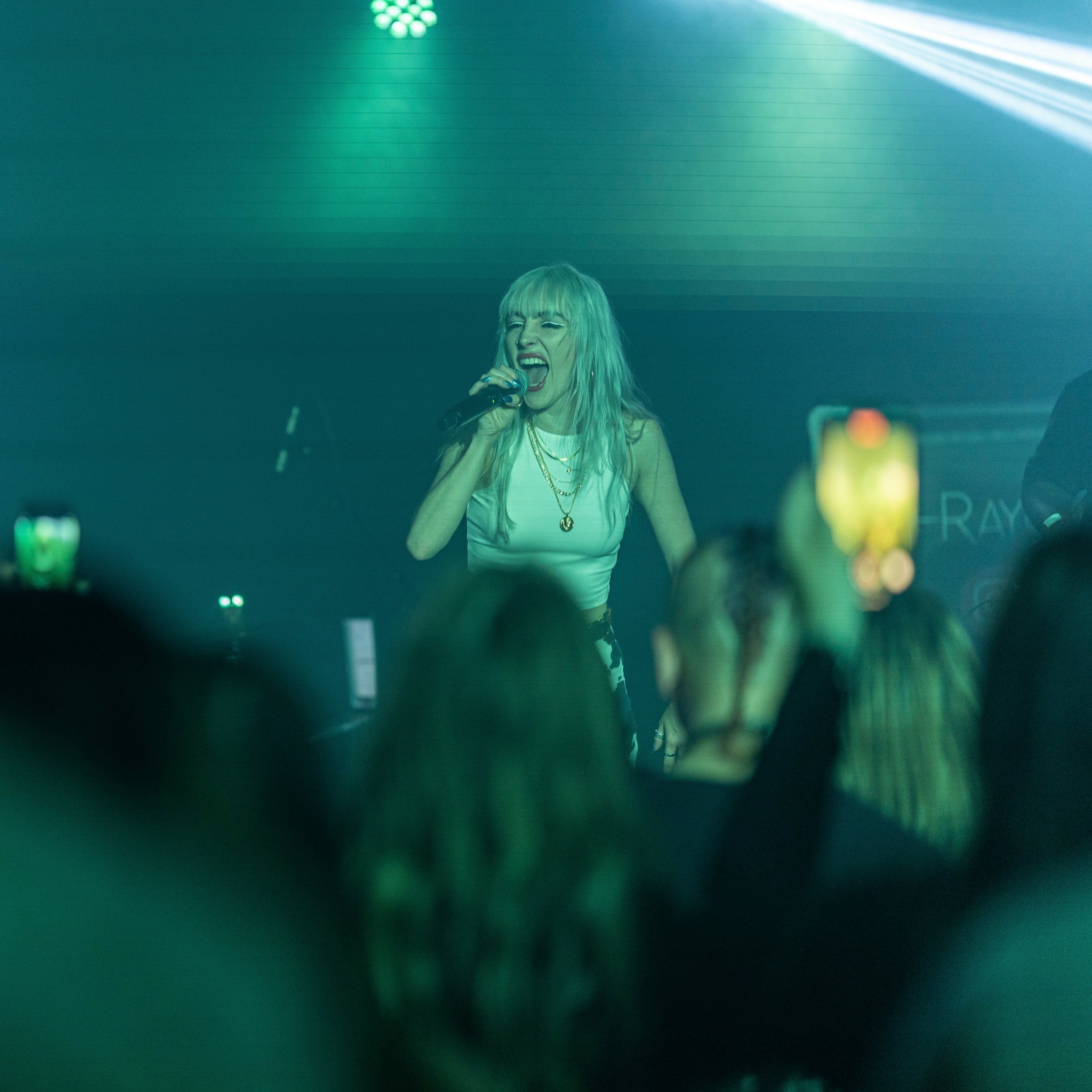 Female singer performing on stage with audience in foreground, green and blue lighting, holding microphone, smiling.